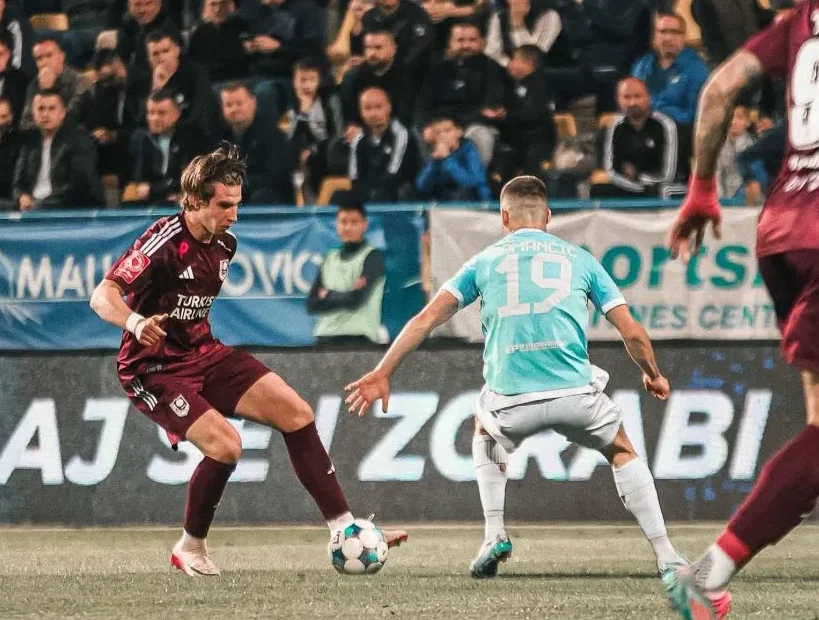 Fudbaleri u bordo i plavoj opremi u borbi za loptu na stadionu, sa publikom u pozadini.