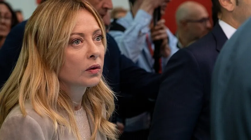 Političarka Giorgia Meloni gleda u stranu sa ozbiljnim izrazom lica dok je okružena drugim ljudima.