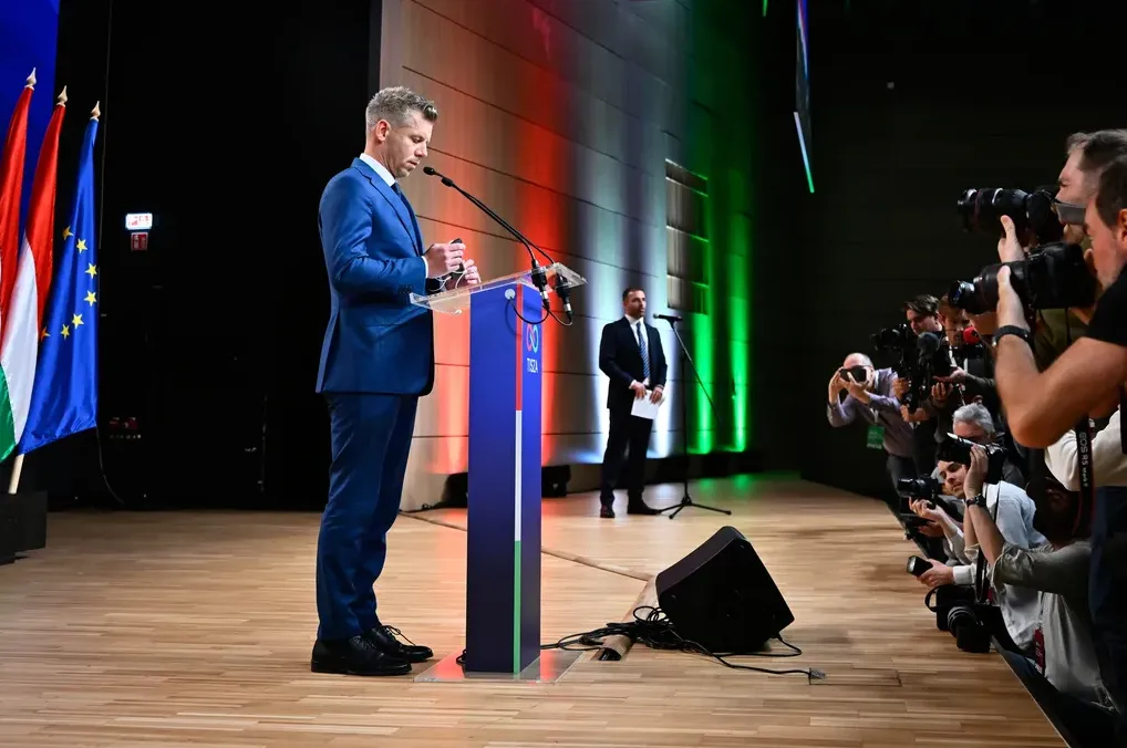 Muškarac u plavom odijelu stoji za govornicom i drži telefon, okružen novinarima sa fotoaparatima.