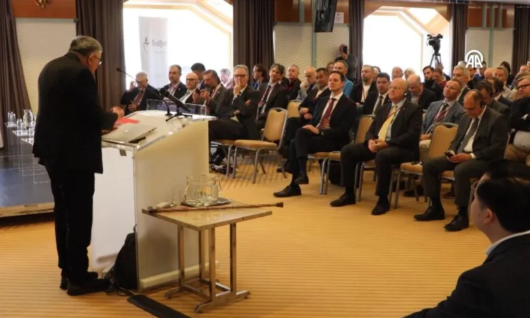 Govornik u crnom odijelu drži govor iza podijuma pred publikom sastavljenom od muškaraca u odijelima.