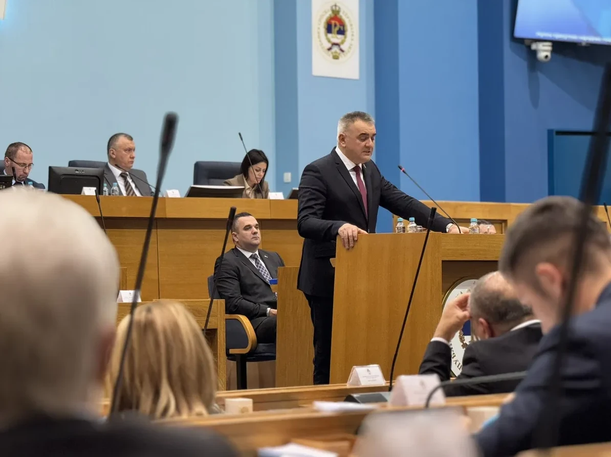 Političar u crnom odijelu govori za govornicom u skupštini, okružen drugim poslanicima.