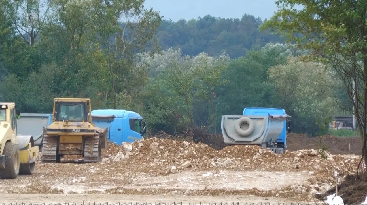 Bager, kamioni i valjak na gradilištu sa šljunkom i drvećem u pozadini.