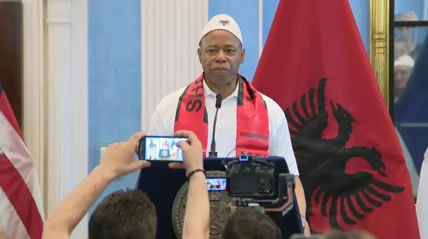 Gradonačelnik Njujorka, Eric Adams, nosi bijelu kapu i crveni šal sa natpisom "SHQIPËRIA" dok stoji ispred albanske zastave.