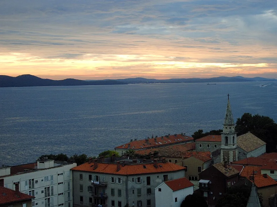 Panoramski pogled na gradski pejzaž pri izlasku sunca, s karakterističnim zvonikom crkve i plavetnilom mora u pozadini.