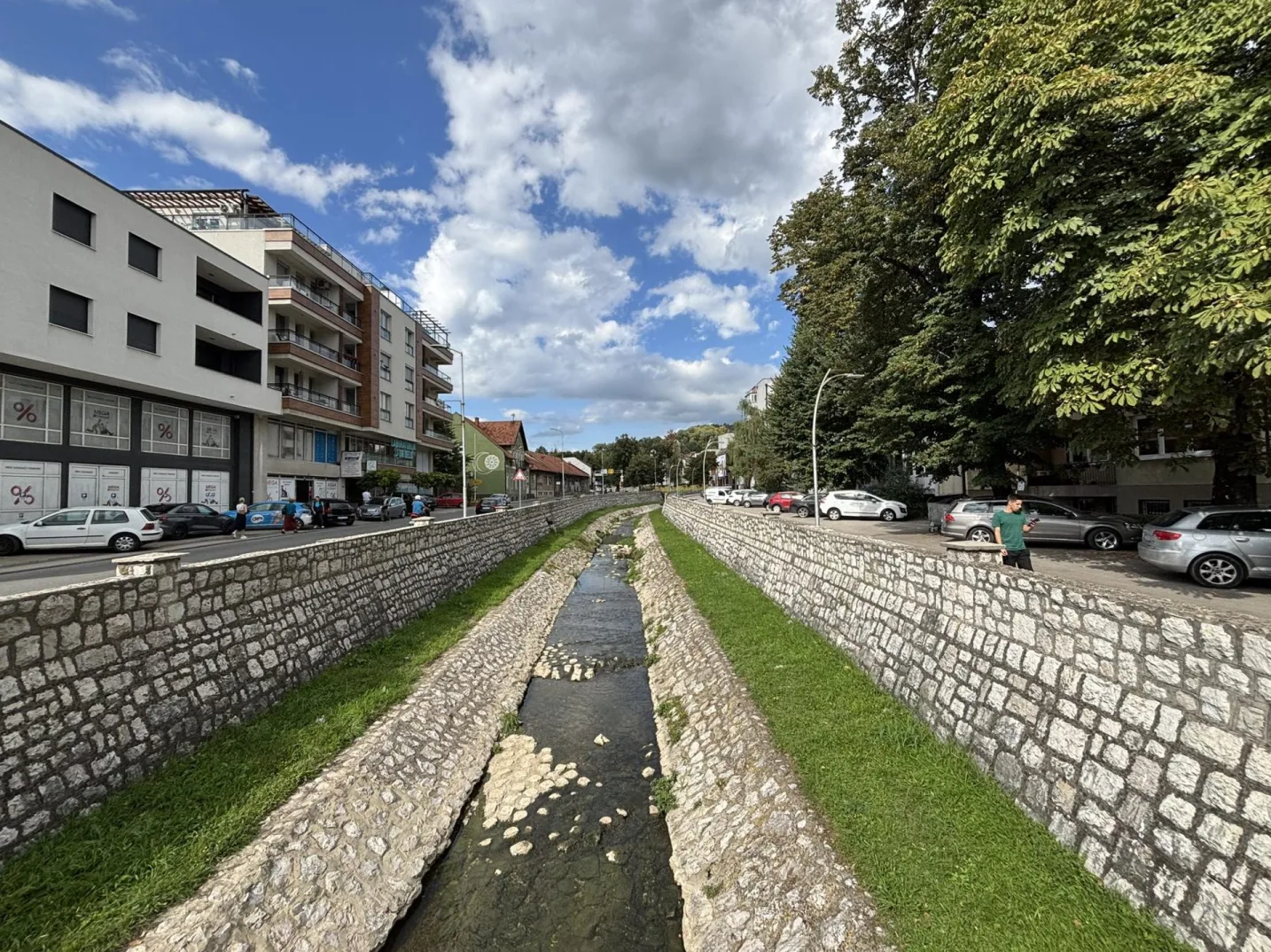Fotografija prikazuje gradski potok sa obostranim kamenim zidanim koritom, okružen zelenom travom i drvećem.