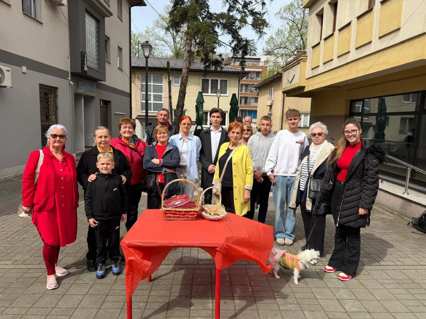 Grupa ljudi, uključujući starije žene, mlade ljude i jedno dijete, stoji ispred stola prekrivenog crvenim stolnjakom na kojem se nalaze dvije košare sa stvarima. U blizini stola je mali bijeli pas obučen u šareni džemper.