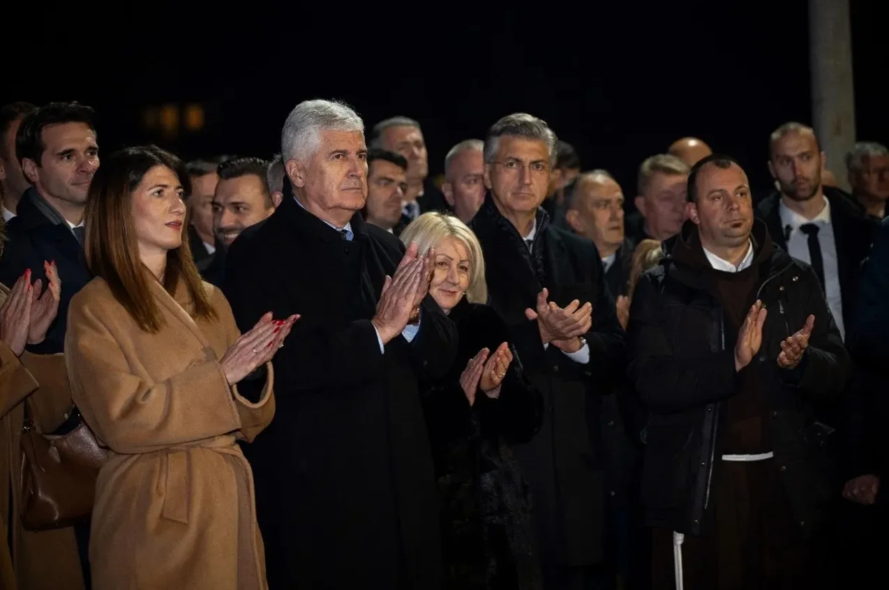 Grupa ljudi, među kojima su muškarci i žene, stoje na otvorenom i aplaudiraju. Neki od prisutnih su poznate ličnosti, sudeći po njihovom ponašanju i pažnji koju su im posvetili ostali okupljeni.
