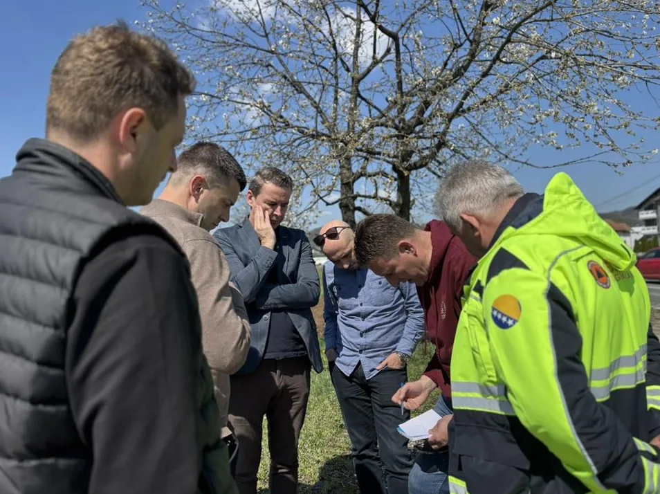 Grupa muškaraca okupljena vani, razgovaraju i gledaju u papire.