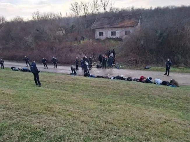 Policija okružuje grupu ljudi koji leže na zemlji u blizini napuštene kuće.