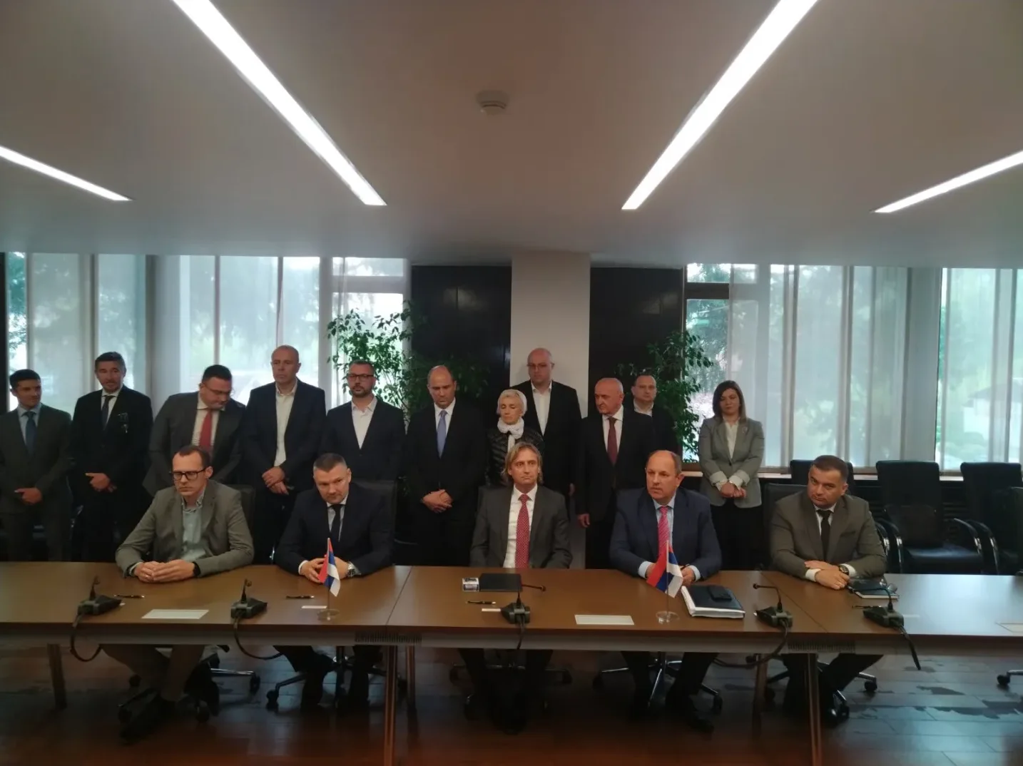 Grupa zvaničnika u svečanim odijelima sjedi i stoji oko velikog stola u moderno opremljenoj dvorani.