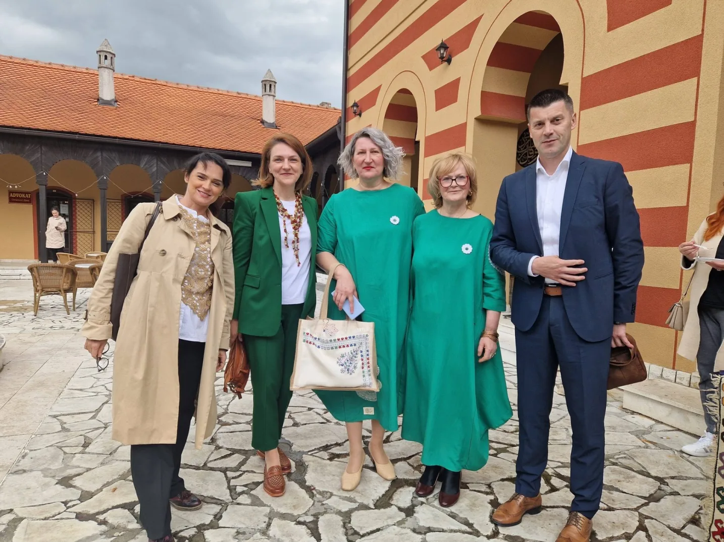 Grupna fotografija pet osoba u elegantnoj odjeći, stoje ispred zgrade sa žutim i crvenim prugama i kamenim popločanjem.