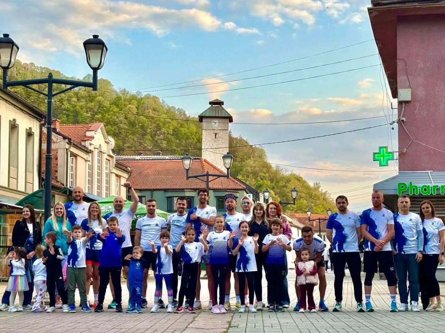 Grupa trkača, među kojima ima i djece, pozira za fotografiju ispred zgrade sa natpisom "Pharmacy" i zelenim znakom apoteke.