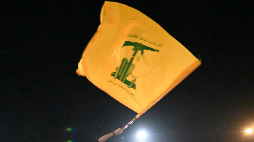 Žuta zastava Hezbolaha sa zelenim logom koji prikazuje pušku AK-47 ukrštenu sa knjigom i sabljom, koja se vijori na noćnom nebu.