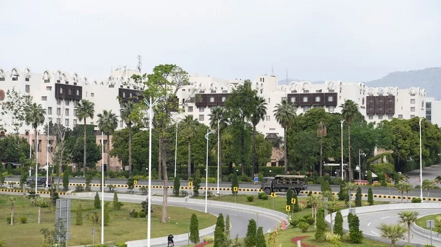 Velika bijela zgrada hotela sa karakterističnim arhitektonskim detaljima i tamnim prozorima, okružena bujnim zelenilom, palmama i drvećem. Ispred zgrade je raskrsnica sa saobraćajnicom i nekoliko saobraćajnih znakova. U jednom smjeru se vidi vojni kamion sa vojnicima, a u drugom smjeru se kreće motociklista. Nebo je oblačno.