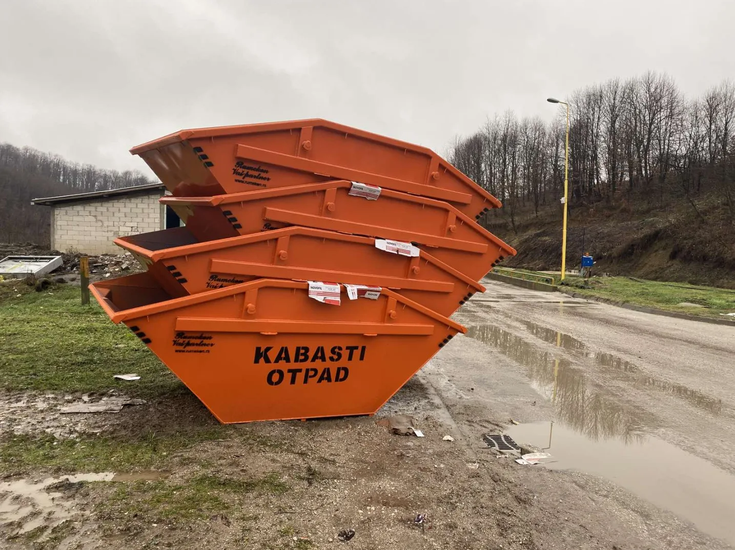 Hrpa narandžastih kontejnera za kabasti otpad parkiranih na blatnjavom putu pored šume i zgrade.
