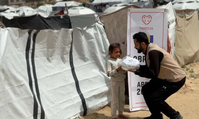 Čovjek u prsluku "Pomozi.ba" daje paket pomoći djevojčici u izbjegličkom kampu.