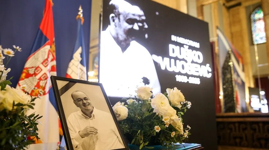 Okvir sa crno-bijelom fotografijom Duška Vujoševića sa buketom bijelog cvijeća u prvom planu, te ekranom sa njegovim likom i imenom te zastavama Srbije u pozadini.