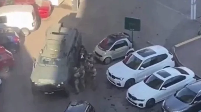 Policijski službenici u opremi okružuju blindirano vozilo parkirano pored nekoliko automobila, uključujući mali Smart automobil i dva bijela BMW-a.