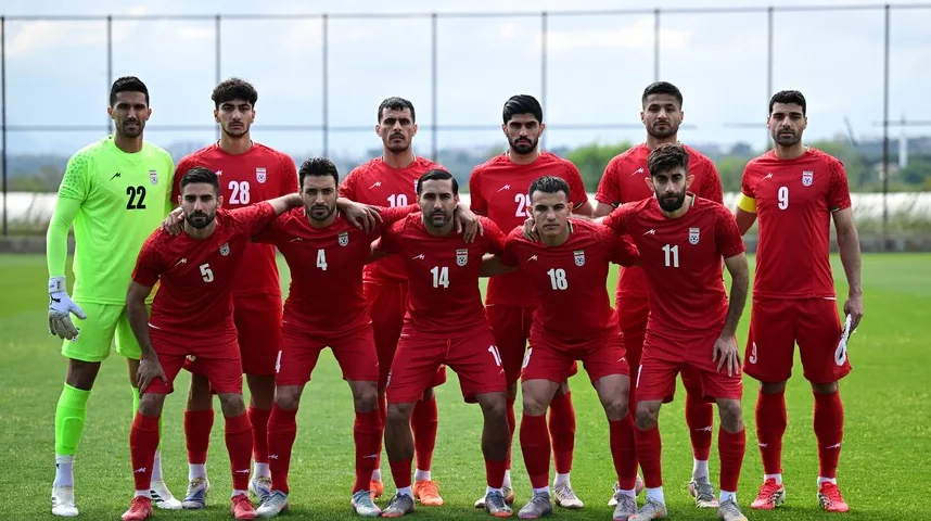 Tim iranske nogometne reprezentacije pozira za fotografiju na zelenom nogometnom terenu. Nogometaši nose crvene dresove i kratke hlače, dok golman nosi zeleni dres.