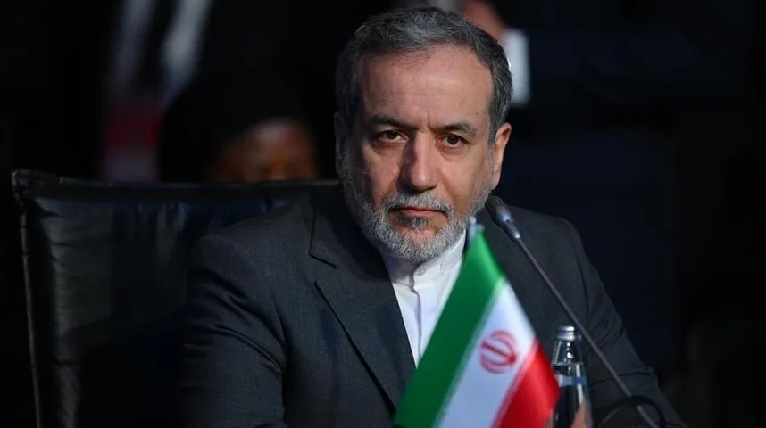 Portret iranskog političara sa bradom i sivom kosom, obučenog u tamno odijelo i bijelu košulju, sa zastavom Irana ispred sebe.