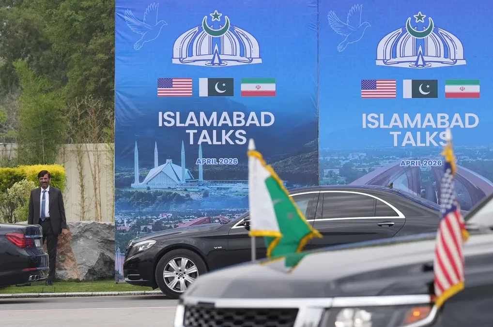 Zvaničnik stoji pored crnog automobila ispred velikog plakata na kojem piše "Islamabad Talks April 2026" sa zastavama SAD-a, Pakistana i Irana.