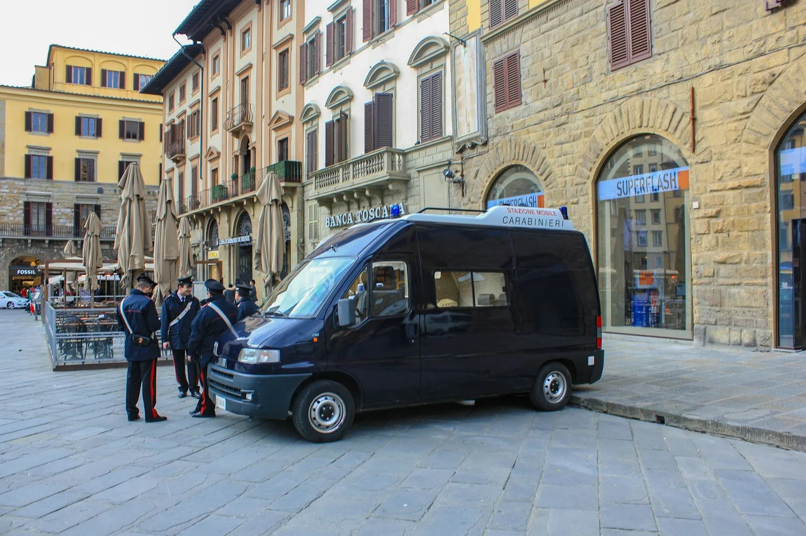 Italijanski karabinjeri u uniformama stoje pored tamnog kombija marke Fiat Ducato s oznakama "Carabinieri" i "Stazione Mobile" na krovu. U pozadini se vide zgrade s arhitekturom tipičnom za Italiju, uključujući natpis "Banca Toscana" i prodavnicu "Superflash".