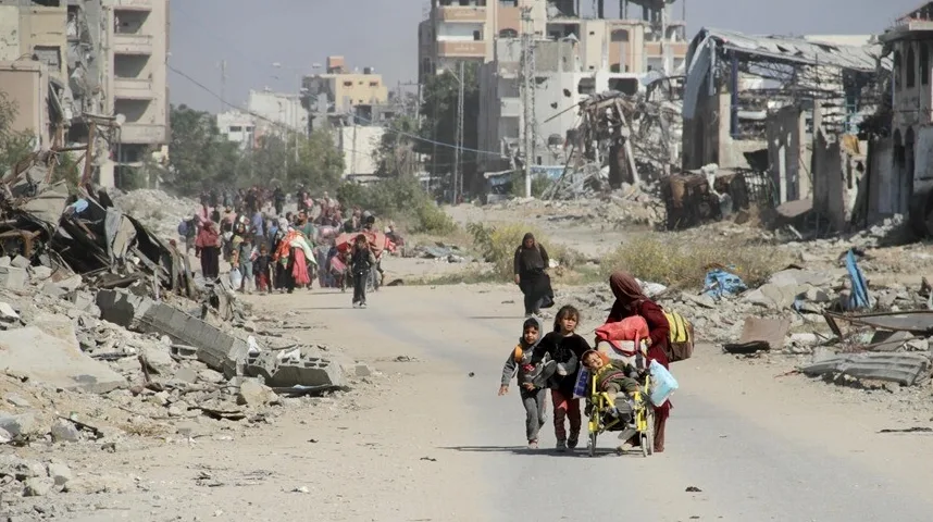 Djeca i odrasli hodaju razrušenom ulicom u ratnom području, neki nose prtljag, dok žena gura dječja kolica.