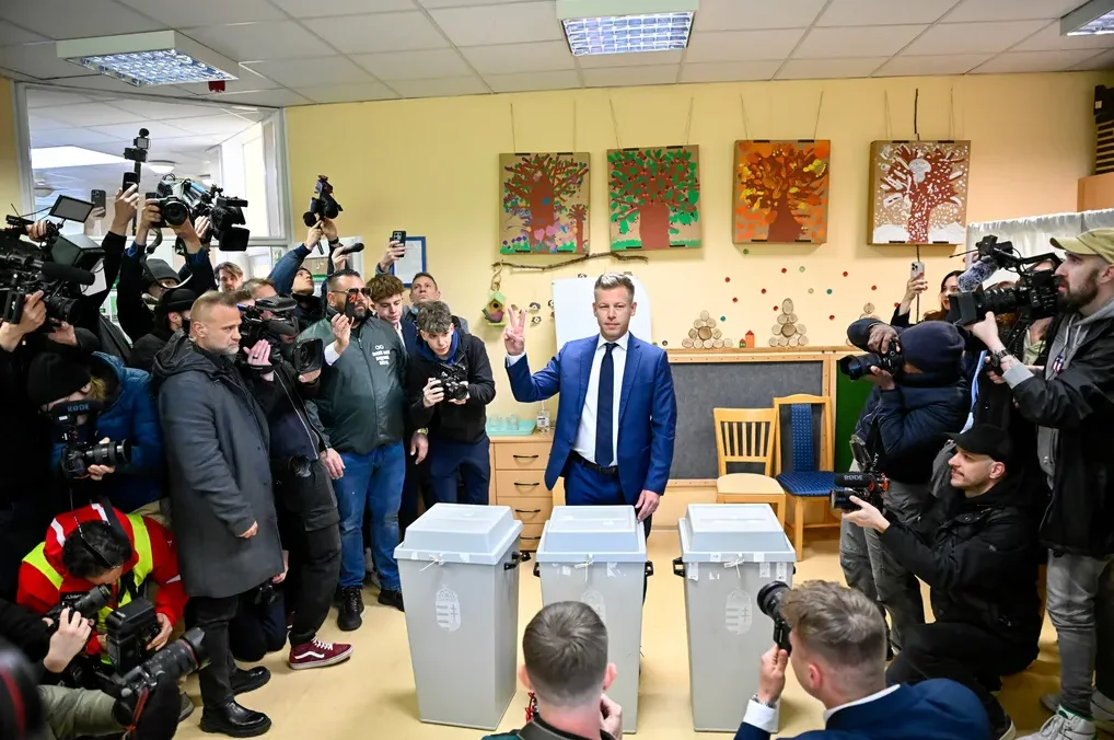 Političar u plavom odijelu diže dva prsta pred biračkim kutijama, okružen velikim brojem novinara s fotoaparatima i kamama.