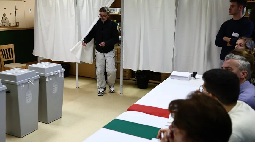 Čovjek izlazi iz glasačke kabine, podižući zastor, dok su u prvom planu glasačke kutije i stol sa ljudima koji čekaju.