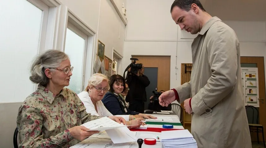 Čovjek u dugom kaputu predaje glasački listić u glasačkoj kabini, dok ga promatraju članovi biračkog odbora i medijske ekipe.