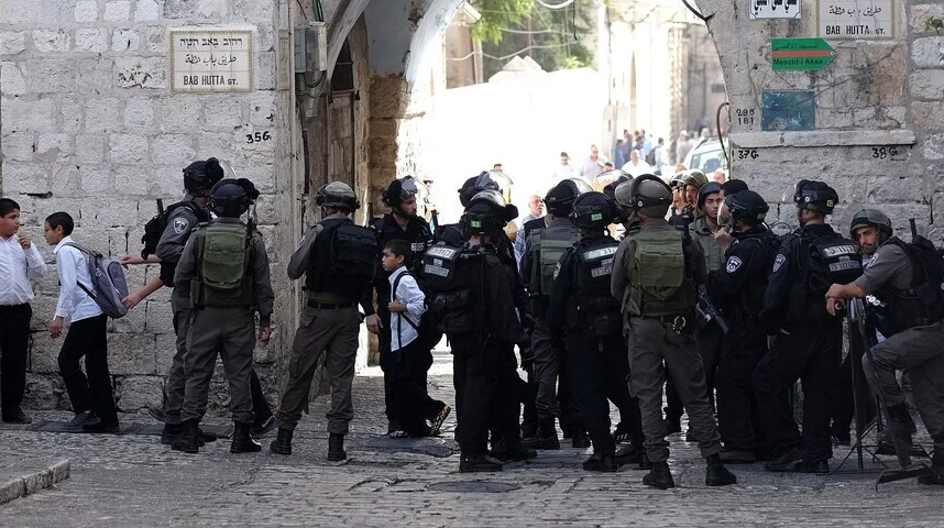 Izraelski policajci u punoj opremi okružuju grupu dječaka u bijelim košuljama i crnim pantalonama, od kojih jedan nosi ranac, na ulici popločanoj kamenom. Iznad njih se nalaze natpisi na hebrejskom i arapskom jeziku, uključujući i oznaku "Bab Hutta St."