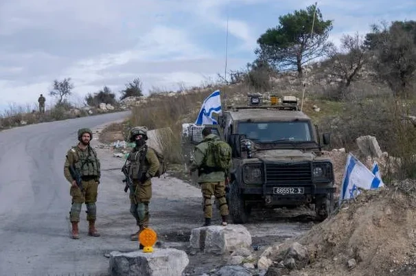 Dva izraelska vojnika u punoj ratnoj opremi stoje na ivici puta, ispred njih se nalazi vojno vozilo sa izraelskom zastavom, a u pozadini se vidi brdoviti teren sa rastinjem.