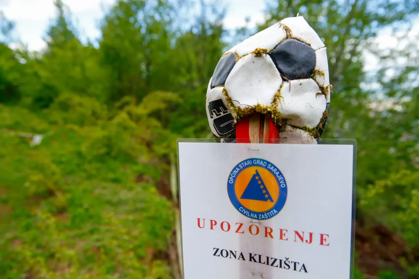 Oštećena lopta za fudbal s mahovinom vidljivom na šavovima, postavljena na vrhu znaka upozorenja za klizište.