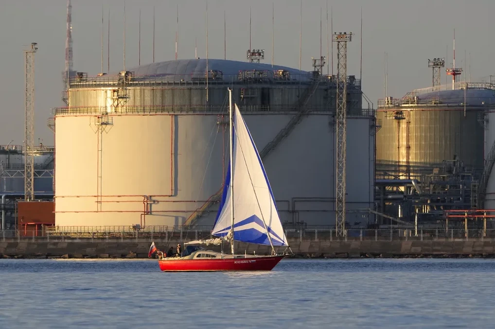 Crveni jedrenjak s bijelim jedrima plovi mirnim plavim morem ispred velikih industrijskih rezervoara u pozadini.