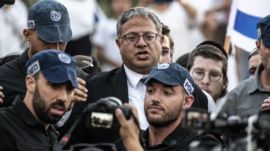 Jevrejski političar sa naočalama i crnim sakoom, okružen muškarcima u tamnim majicama i kačketima sa policijskim oznakama, dok ga fotografi snimaju.