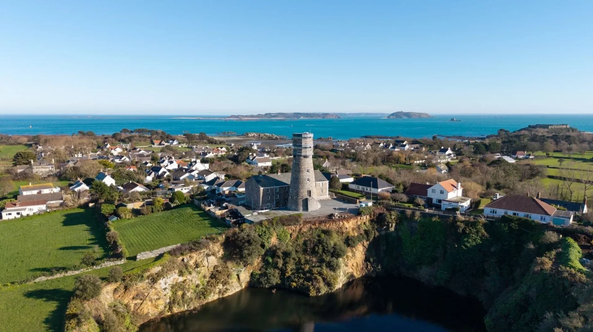 Pogled iz zraka na kameni toranj sa višespratnom strukturom, okružen malim naseljima bijelih kuća i zelenim poljima, sa tirkiznim morem i ostrvima u daljini.