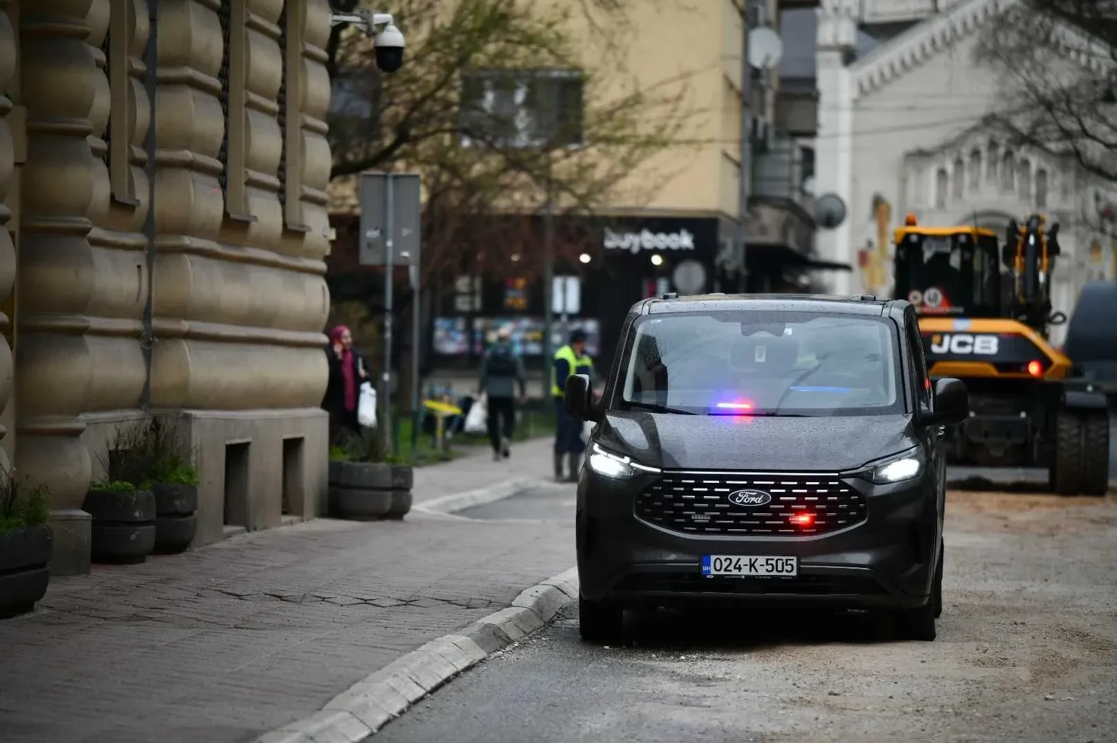 Tamni Ford kombi sa uključenim plavim i crvenim rotacionim svjetlima, parkiran na ulici sa desne strane kadra. U pozadini se vidi žuti bager marke JCB i zgrada sa fasadom.
