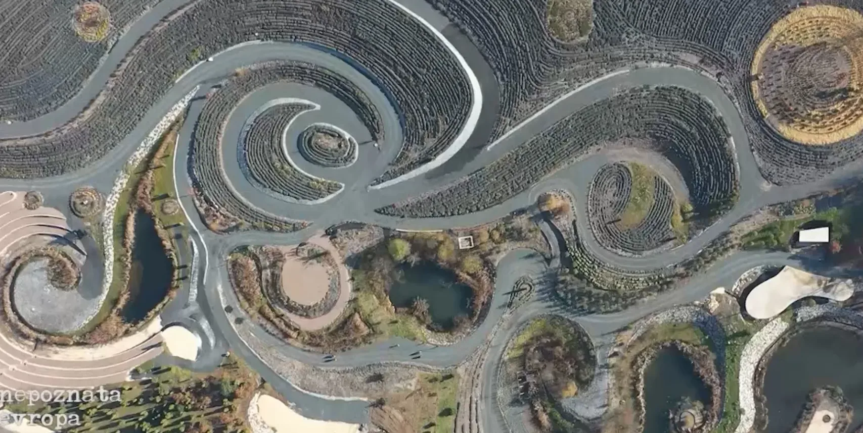 Ptičija perspektiva na kompleks koji prikazuje krivudave staze, vodene površine, terasaste vrtove i kružne obrasce zasađenih biljaka.