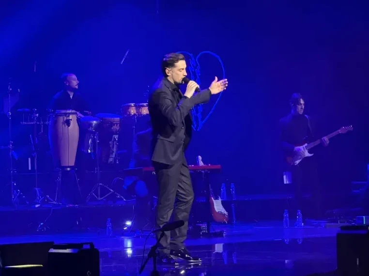 Muškarac u crnom odijelu pjeva na sceni, osvijetljen plavim svjetlom. U pozadini bend svira instrumente, uključujući bubnjeve i gitaru. Na ekranu iza njih projektovan je stilizovani srcoliki oblik.