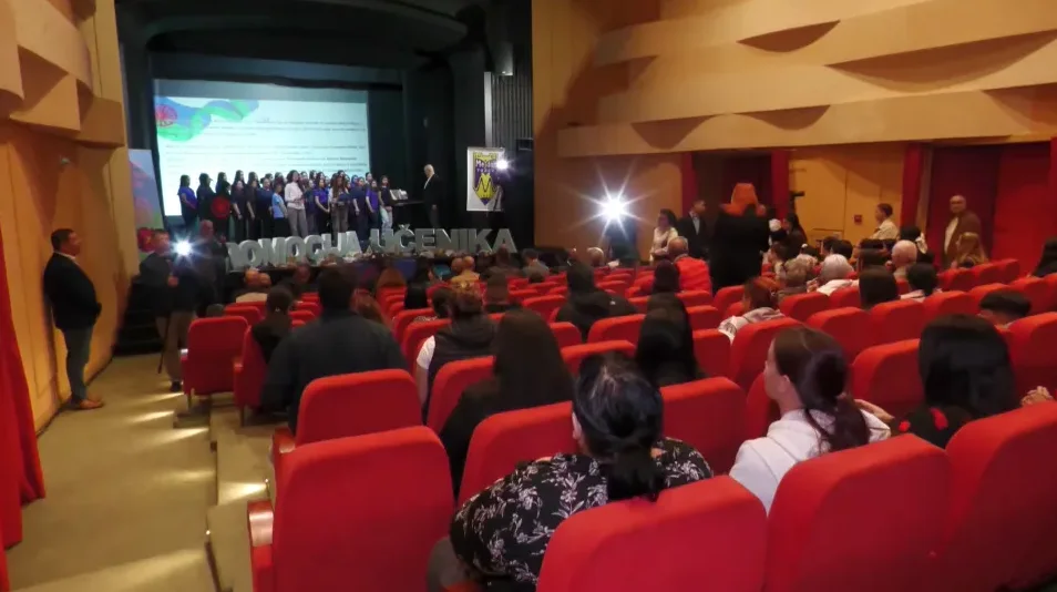 Grupa pomagača učenika stoji na pozornici ispred velikog ekrana i publike u crvenim sjedištima.