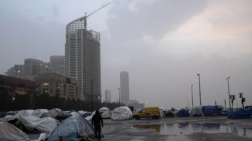 Skupina šatora i improvizovanih skloništa podignuta na parkingu ispred modernih zgrada i gradilišta, s jednim žutim kombijem koji stoji parkiran.