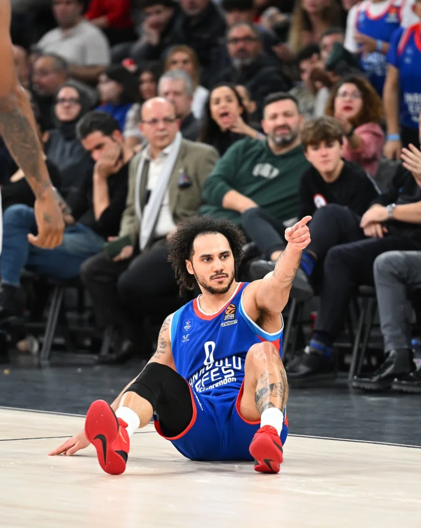 Košarkaš turskog tima Anadolu Efes sa brojem 0 na dresu, u plavom šortsu i crvenim patikama, sjedi na parketu dvorane i pokazuje prstom prema naprijed.