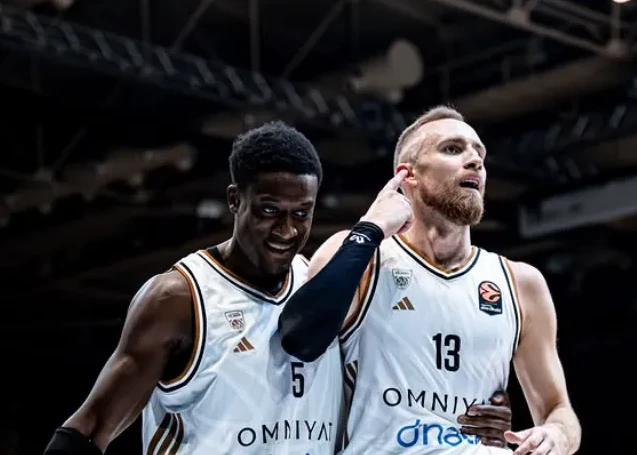 Dva košarkaša u bijelim dresovima, jedan crnac sa brojem 5 i jedan bjelac sa brojem 13, gledaju prema naprijed.