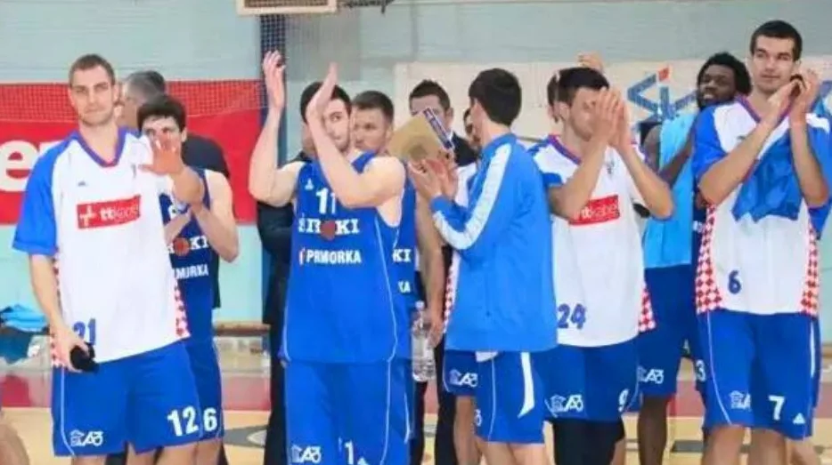 Grupa košarkaša u plavim i bijelim dresovima sa brojevima, neki podižu ruke u znak pobjede ili pozdrava.