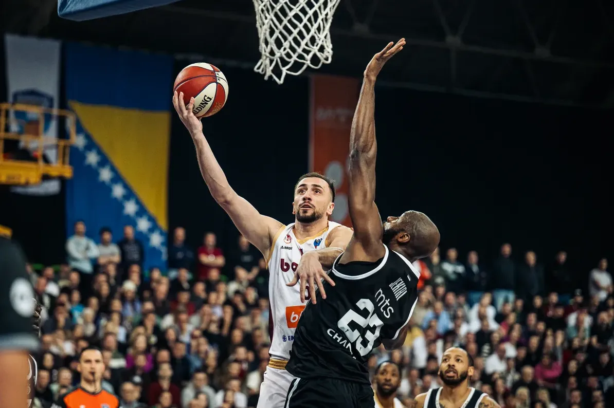 Košarkaš u bijelom dresu sa brojem 9 pokušava položiti loptu u koš dok ga sprječava tamnoputi košarkaš u crnom dresu sa brojem 23.