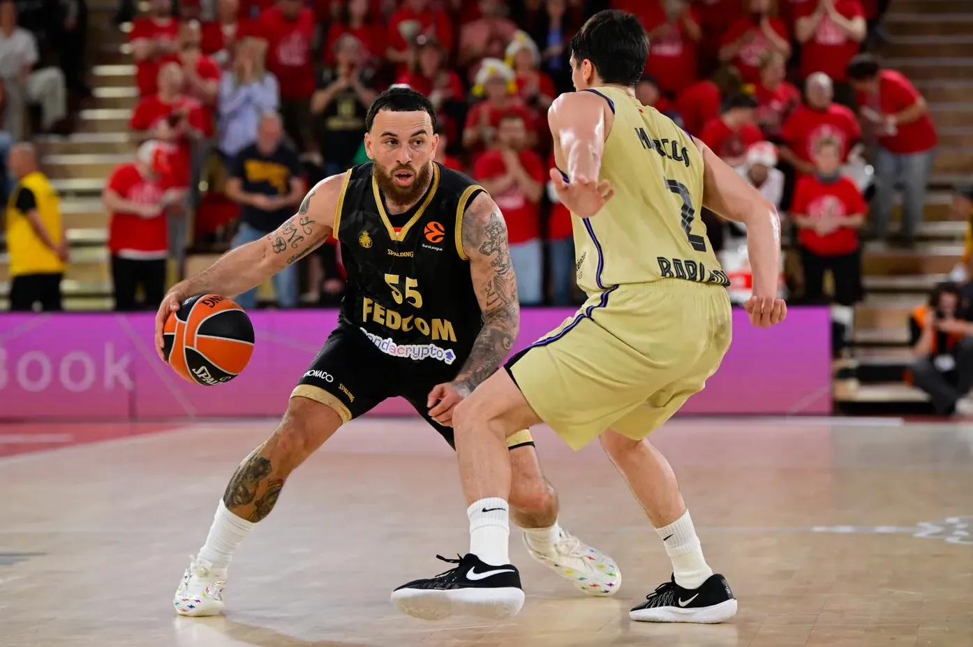 Košarkaš u crnom dresu sa brojem 55, AM Monaco, dribla loptu dok ga čuva igrač u žutom dresu Barcelone.