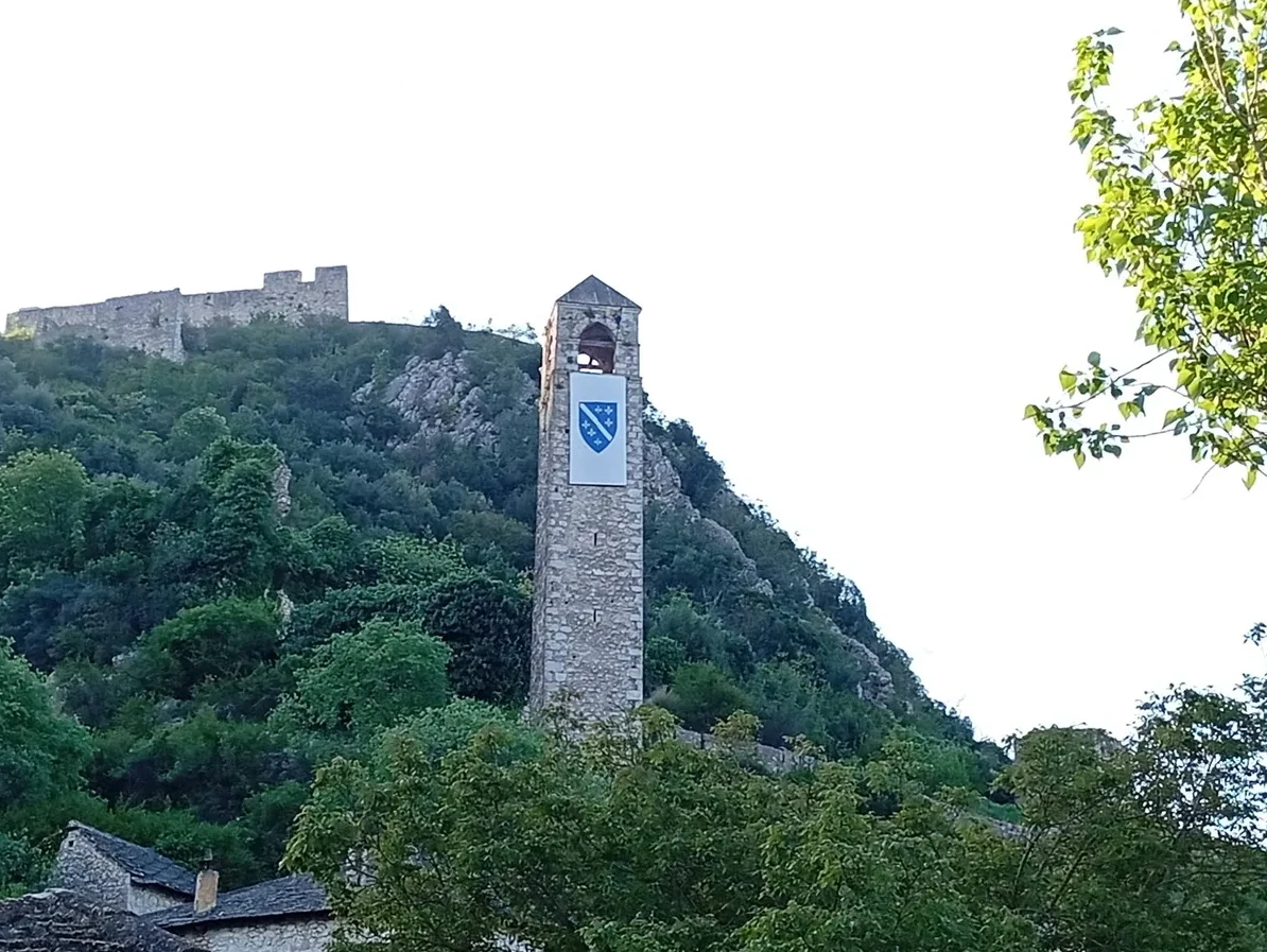 Kamena kula sa zastavom Kraljevine Bosne i ostaci srednjovjekovne gradine iznad nje, okruženi zelenilom.