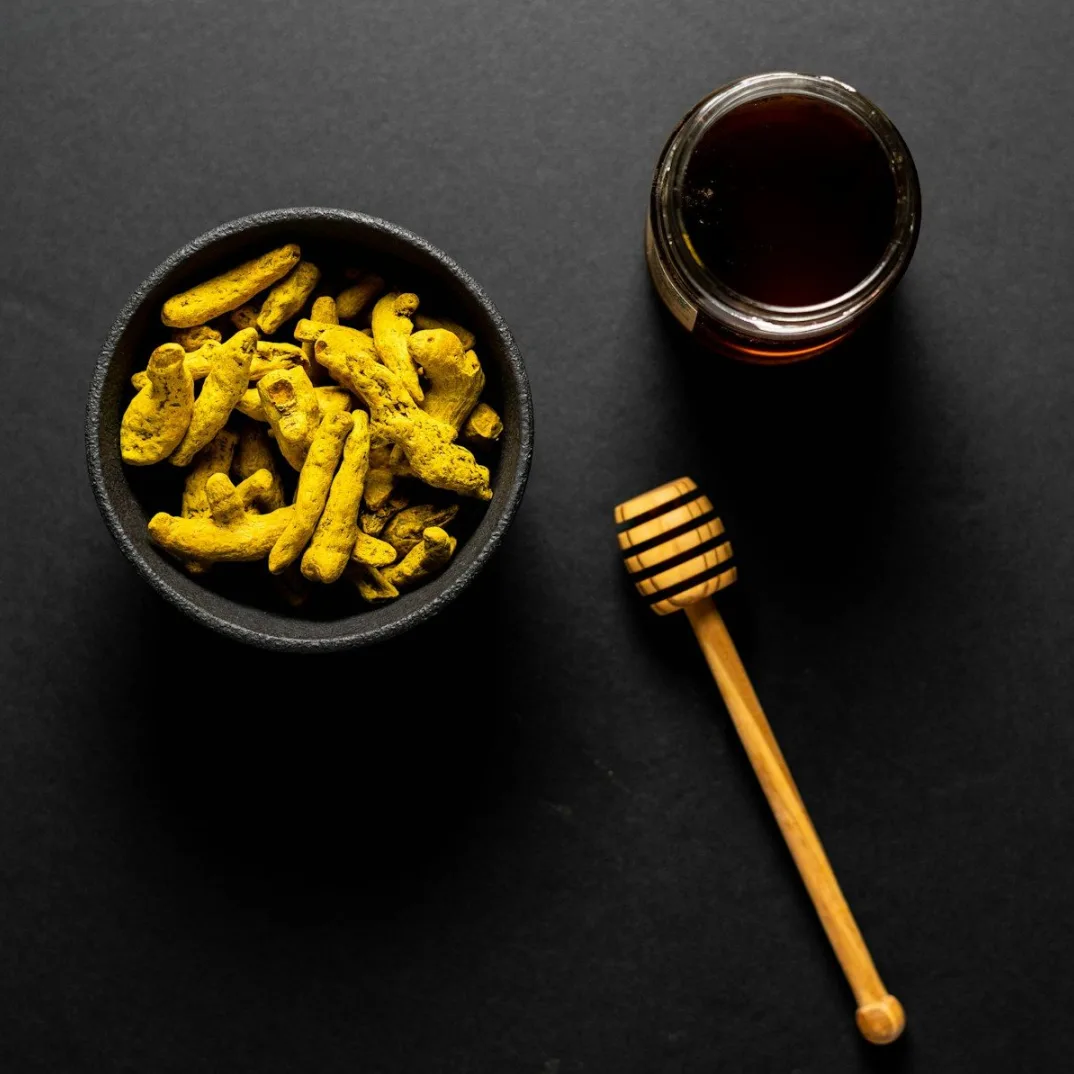 Zdjela punjena suhom kurkumom pored tegle meda i drvenog cjedila za med na crnoj pozadini.