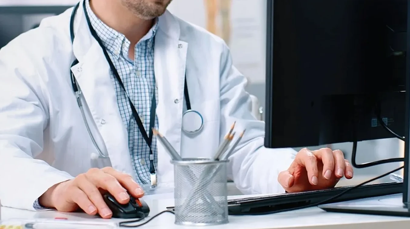Ljekar u bijelom mantilu sa stetoskopom oko vrata koristi kompjuter, pritiskajući miša.