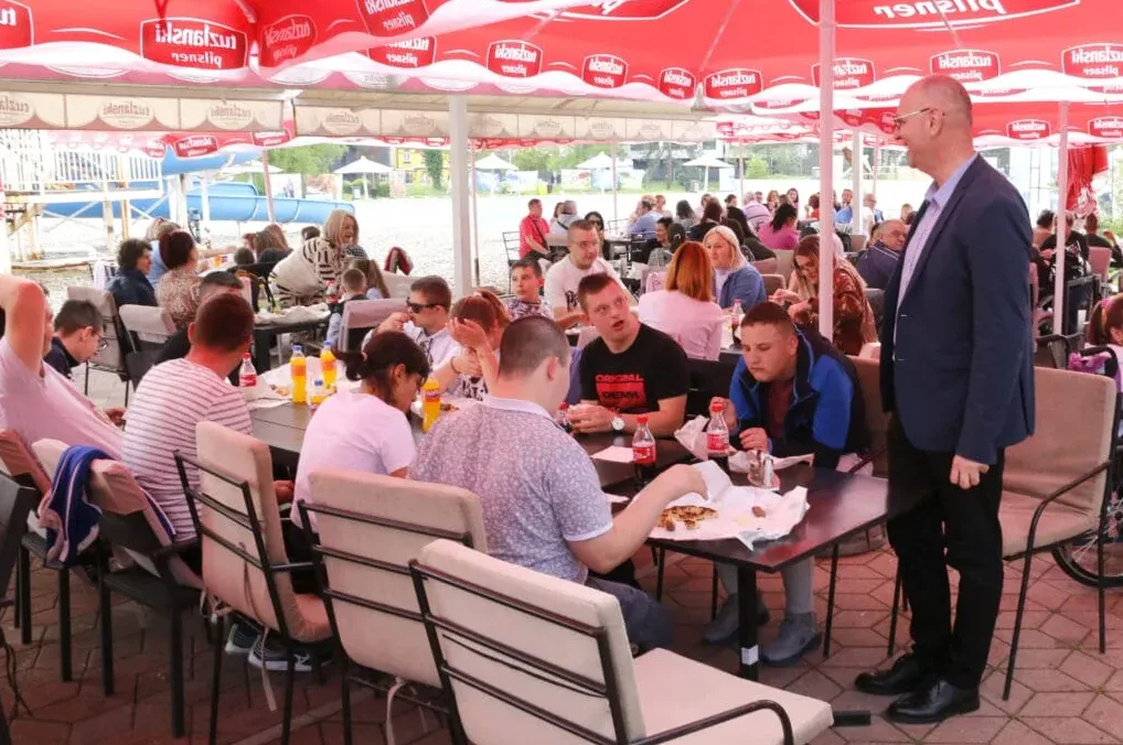 Grupa ljudi sjedi za stolovima u restoranu na otvorenom, ispod crvenih suncobrana. Neki jedu i piju, dok jedan muškarac stoji pored stolova.